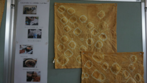 薄茶色の絞り染めのハンカチが2枚、制作の様子の写真と一緒に展示されている写真