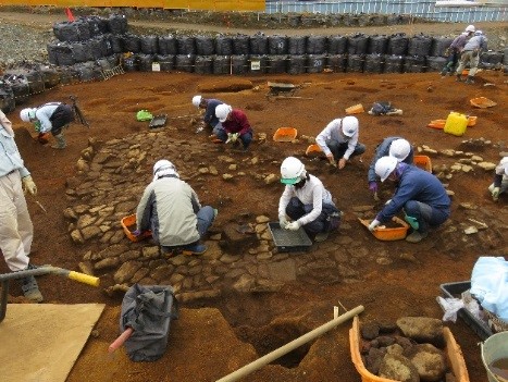 屋外の発掘現場で、複数の作業員がスコップやブラシを使い掘り進めている様子の写真