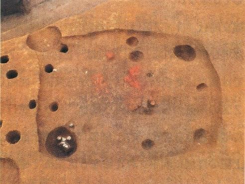 地層が掘り下げられ、地面に複数の穴と赤い模様が点在する、竪穴住居の跡とみられる遺跡を上空から撮影した写真