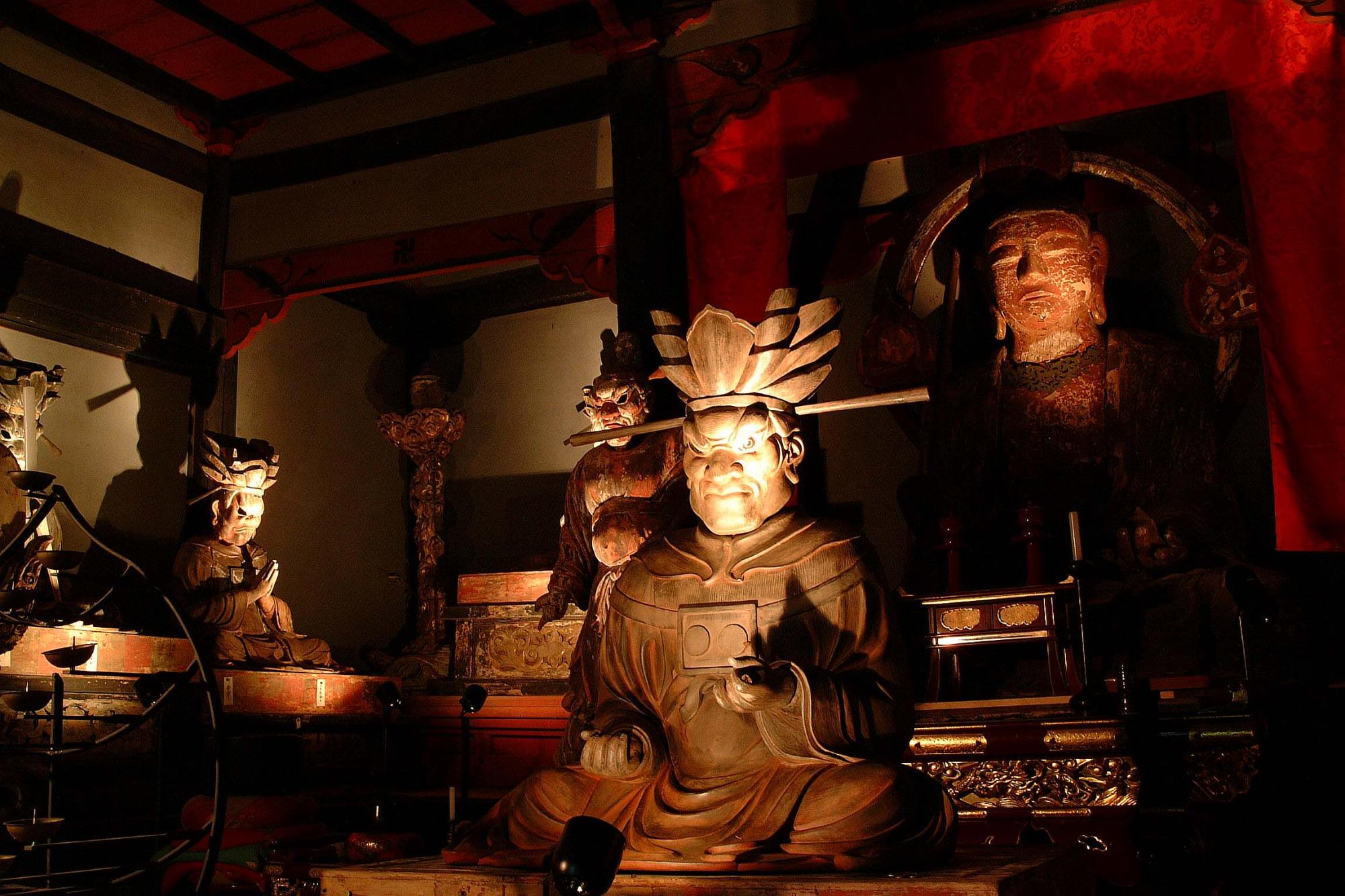 荘厳な寺院の薄暗い空間に、光を浴びた複数の仏像が鎮座し、中央の仏像が正面を睨みつけている写真