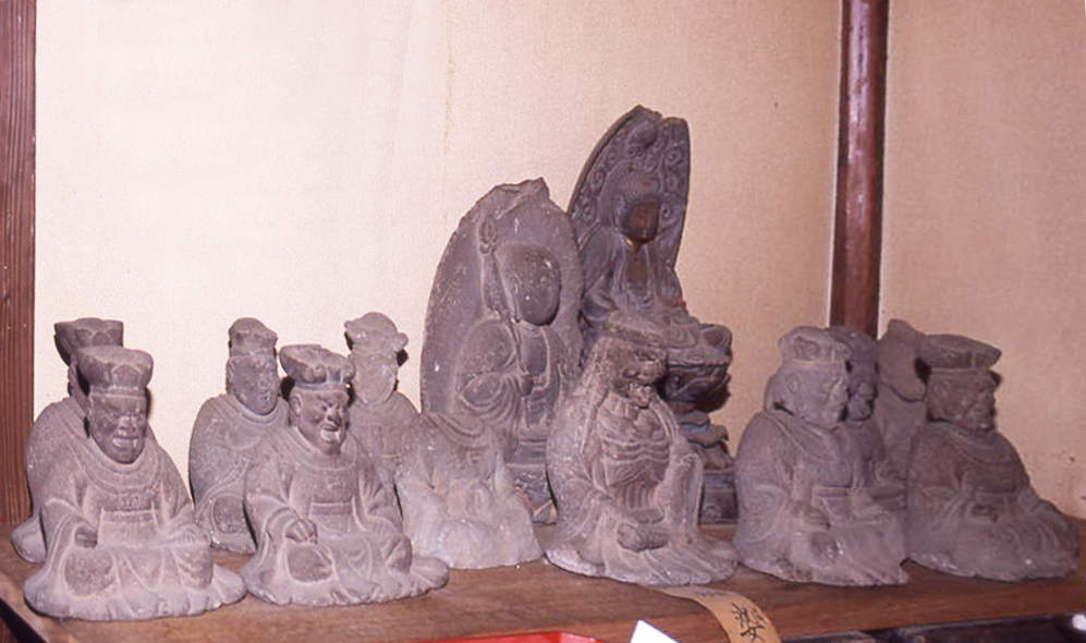 灰色の石造りの複数の坐像が、背景の茶色の壁と柱を背にして、古びた棚の上に並べられている写真