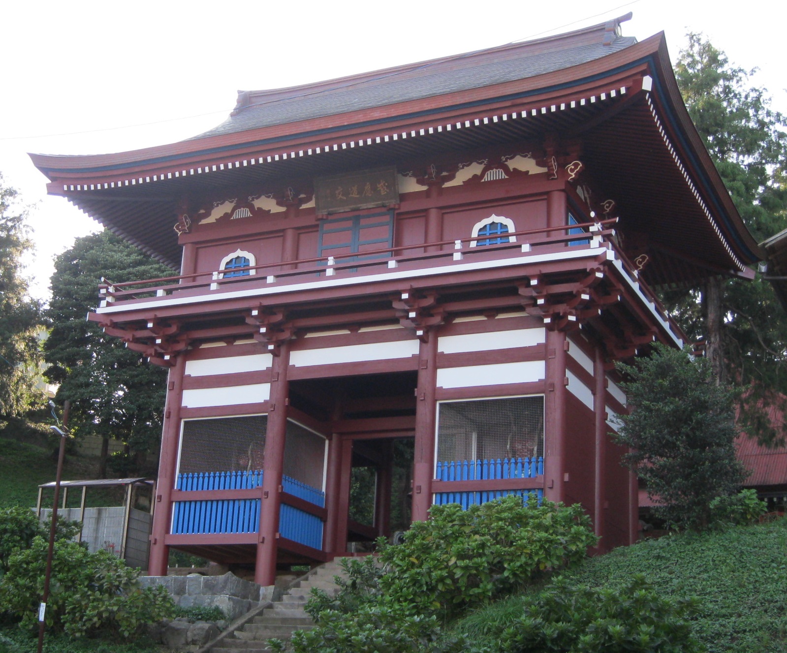 日本風の朱色と白で彩られた二階建ての薬師堂山門が、木々に囲まれた石段の上に建っている様子を捉えた写真