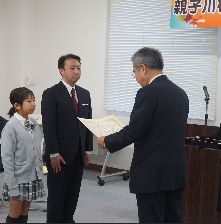 グレーの服を着た女の子と黒いスーツを着た男性が並んで立ち、スーツを着た男性から表彰状を受け取っている写真