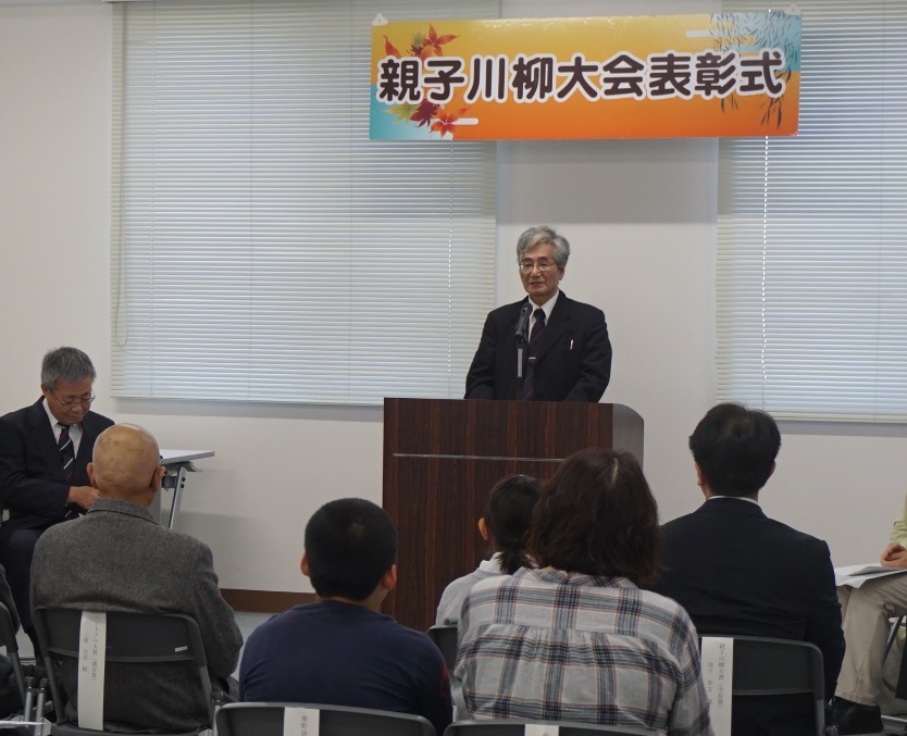 親子川柳大会表彰式と書かれた横断幕の下で、スーツを着た鍛冶実行委員兼審査委員長が、演台に立ち参加者に向かって話している写真