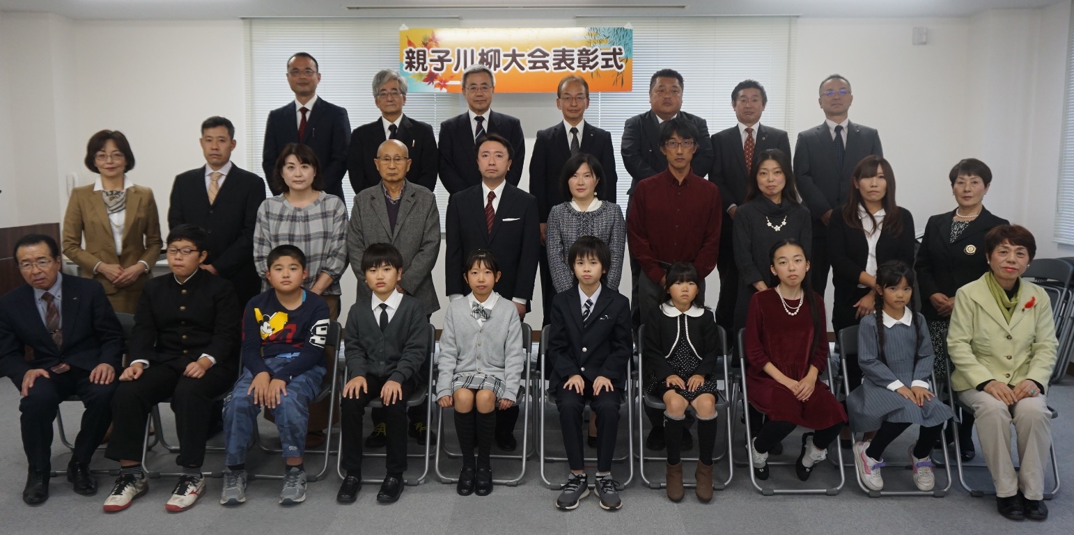 親子川柳大会表彰式と書かれた横断幕の前で、前列の椅子に入賞者の子どもたちが座り、その後ろに保護者や後援団体の方々が立ち3列に並んで写した記念写真