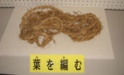 「葉を編む」と書かれた札が置かれ、何本もの藁を束ねて、太く丈夫な縄のような形に編んだものが台の上に置かれている写真