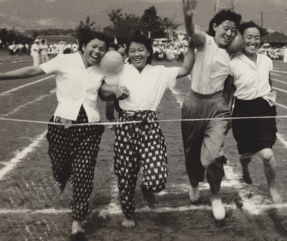 2人一組の白のシャツに柄物のモンペを穿いた女性たちが顔の間にボールを挟んで、笑顔で一斉にゴールテープを切っている、昭和時代の運動会の様子の白黒写真