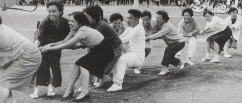 動きやすそうな服装をした男性たちや、着物やミニスカートを履いた女性たちが楽しそうな表情を浮かべ、運動会で綱引きに熱中している様子を写した白黒写真