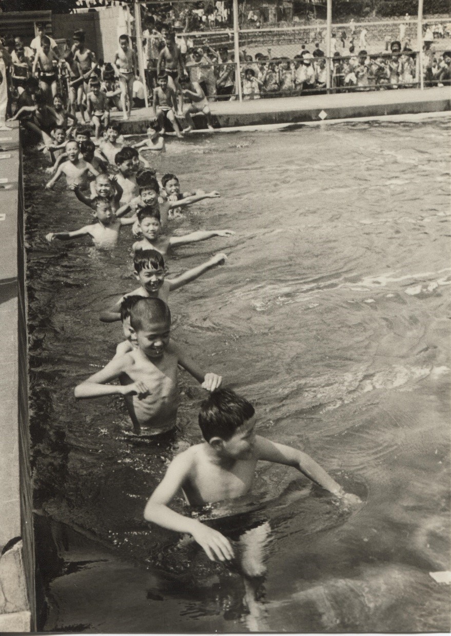 周囲で沢山の子どもたちが見ているなか、楽しそうな表情の子供たちがプールの列に並んでいる様子を、プールサイドから撮影した白黒写真