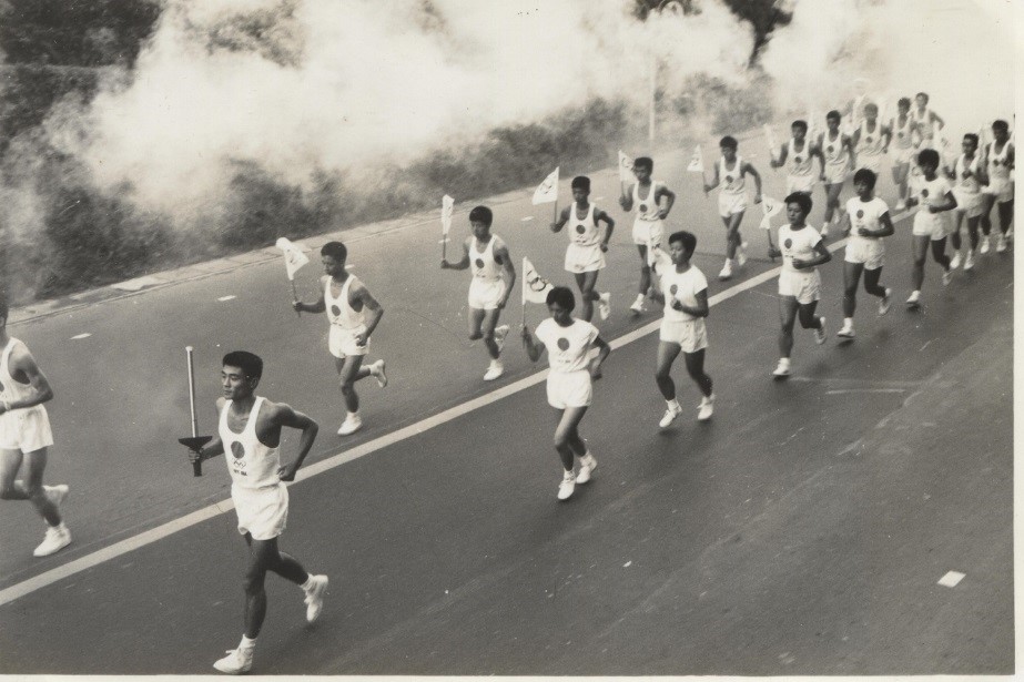 ユニフォーム姿のランナーたちが、煙を伴いながらトーチを掲げ道路を走っているオリンピックの聖火リレーを捉えた白黒写真