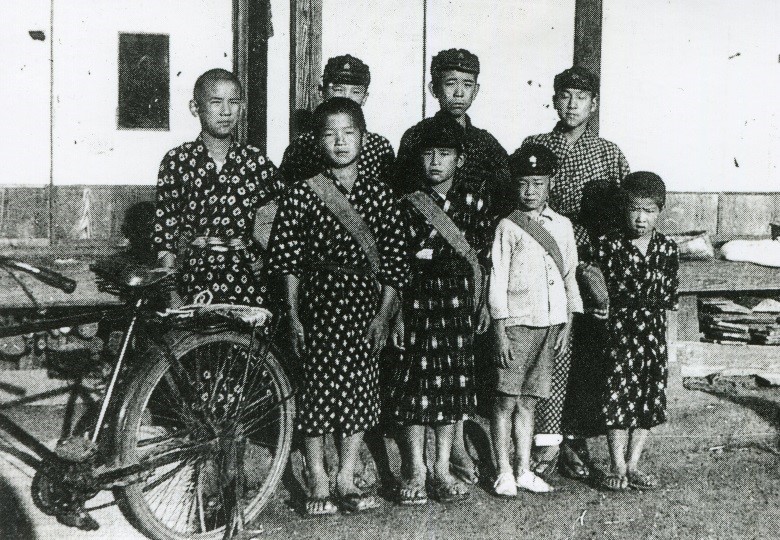 濃い柄の着物や半ズボンなどを着た男の子たちが、建物の前で自転車と共に立ち集合している白黒写真