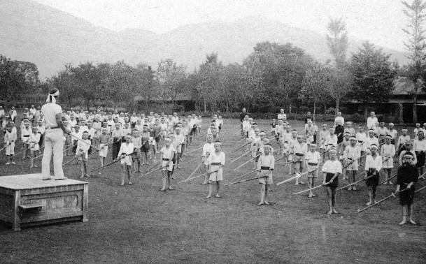 鉢巻きを巻いて体操服を着た大勢の少年たちが、広場に整列して竹刀を構えており、指揮者が一段高い壇上から彼らを指導している様子の白黒写真