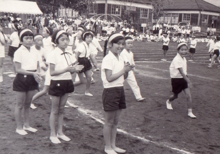 運動会で体操服を着た複数の小学生が、校庭のトラックに並んでおり、楽しそうに拍手をしながら観客や他の生徒の方を向いている白黒写真