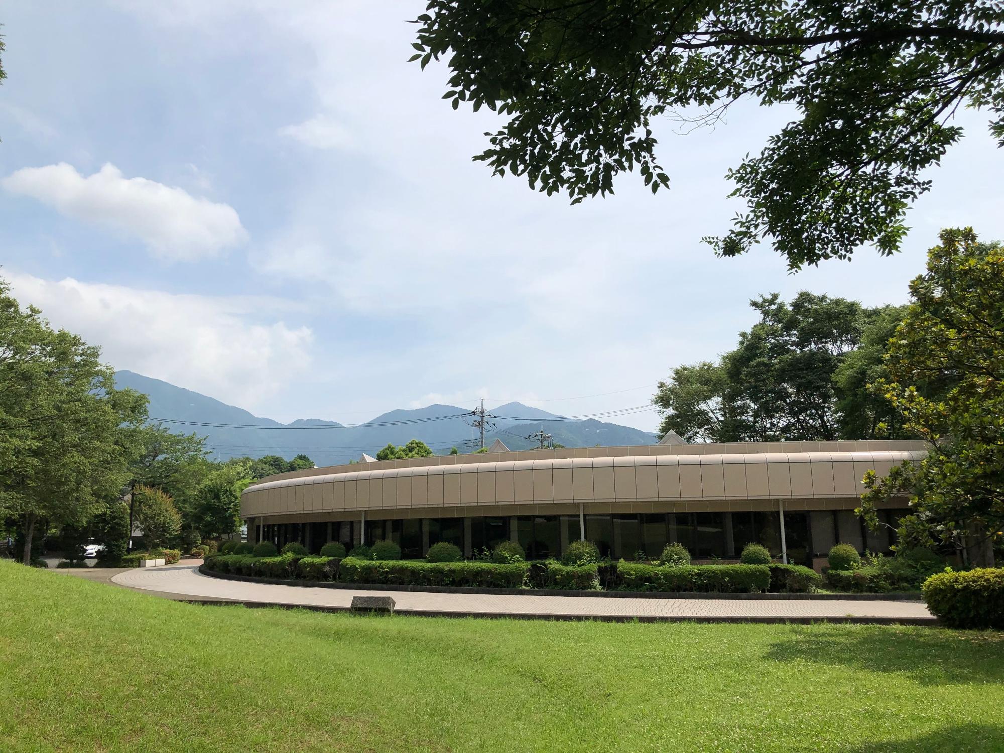 周囲を木々に囲まれ、芝生が広がる古墳の奥に建つ、緩やかな曲線を描いた桜土手古墳展示館の建物の外観写真