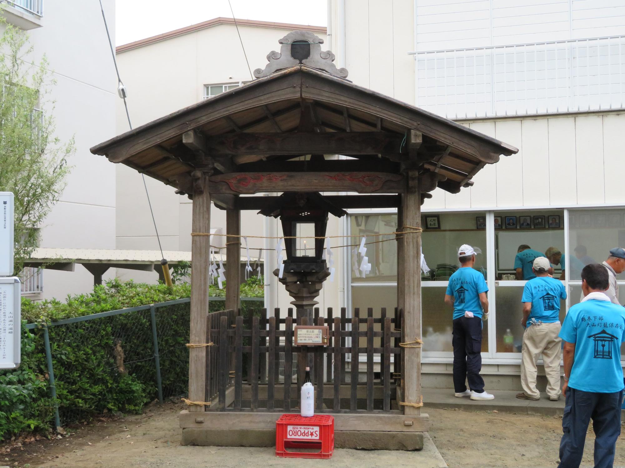 灯籠が設置された覆屋の柱にしめ縄が取り付けられ、周囲には作業を終えた水色のTシャツの男性たちが立っている写真