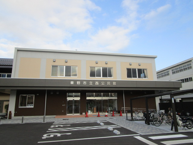 クリーム色と茶色の外観を持つ秦野市立西公民館の建物正面と駐車場を撮影した写真