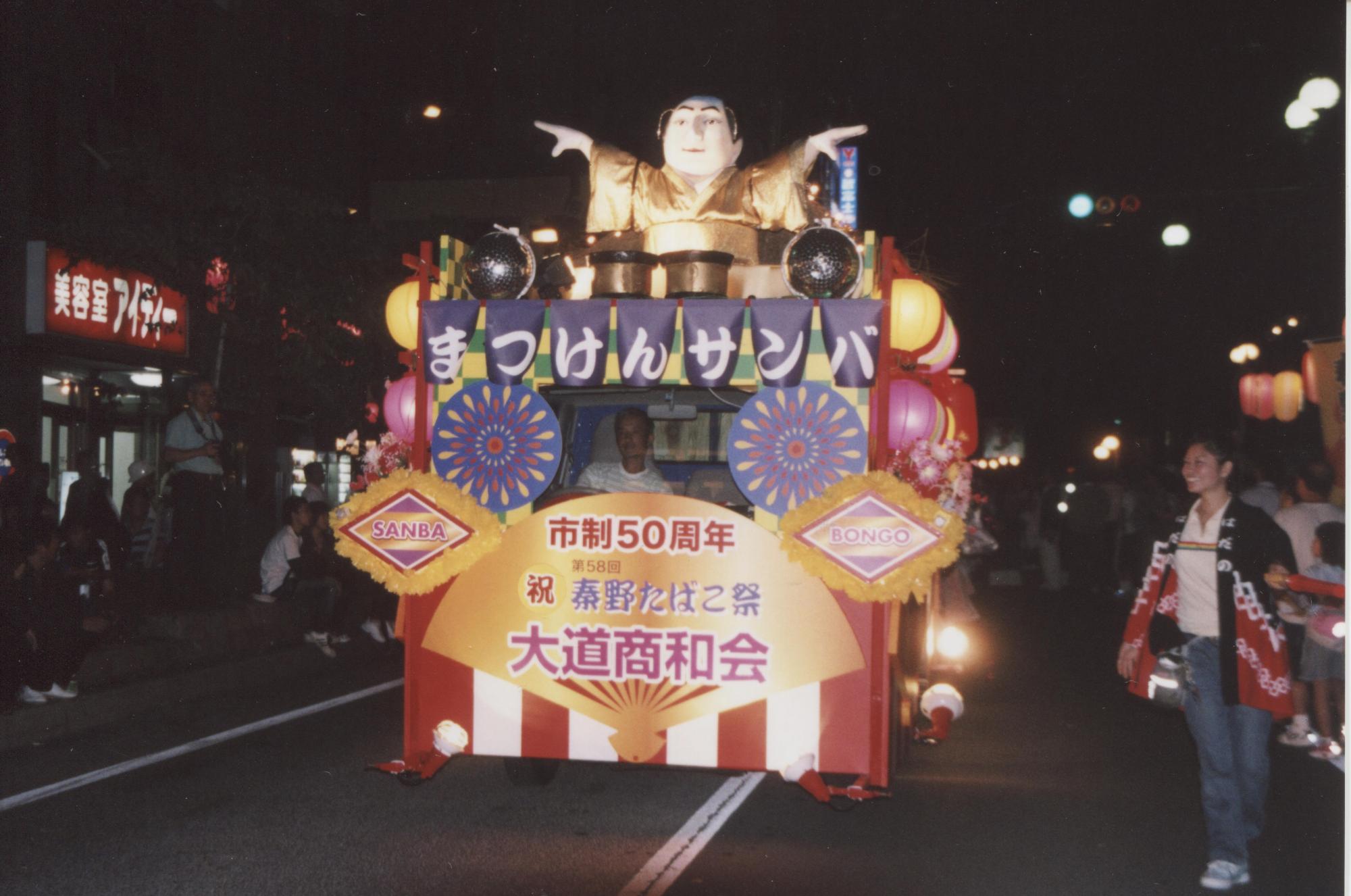 祭りの夜、ライトアップされた山車が道路を進んでおり、その上には両手を広げた人形が飾られ、「まつげんサンバ」「大通商和会」などの文字が見える写真