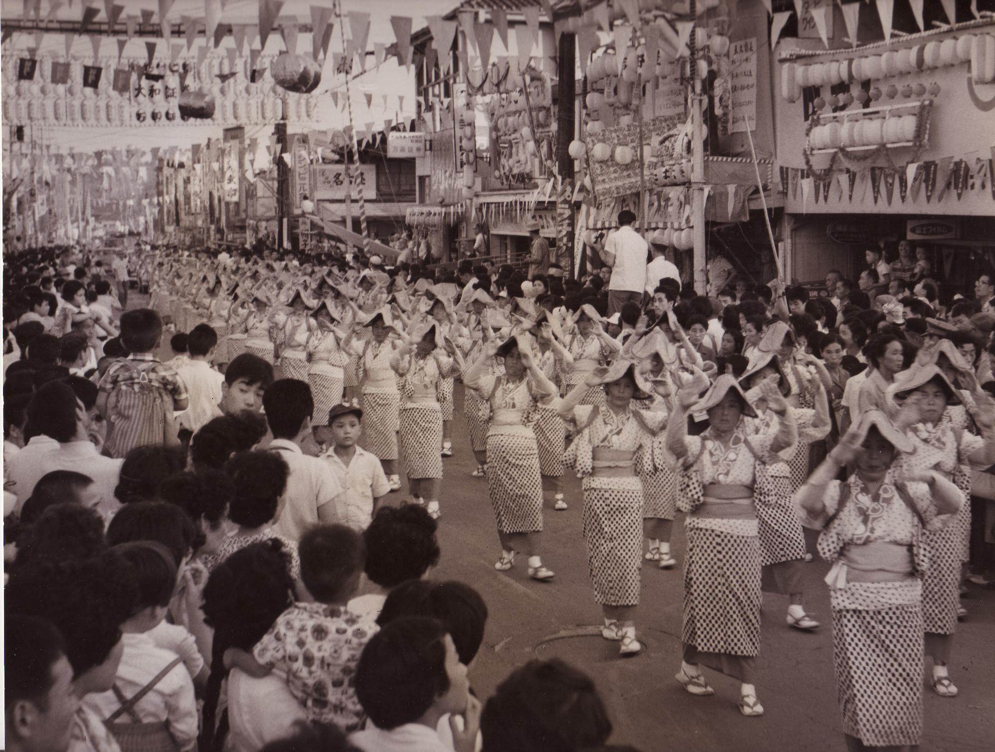 頭に編み笠をかぶり、浴衣を着た大勢の女性が、お囃子に合わせて踊りながらパレードをしており、沿道には多くの観客がいる夏祭りの風景を捉えた白黒写真