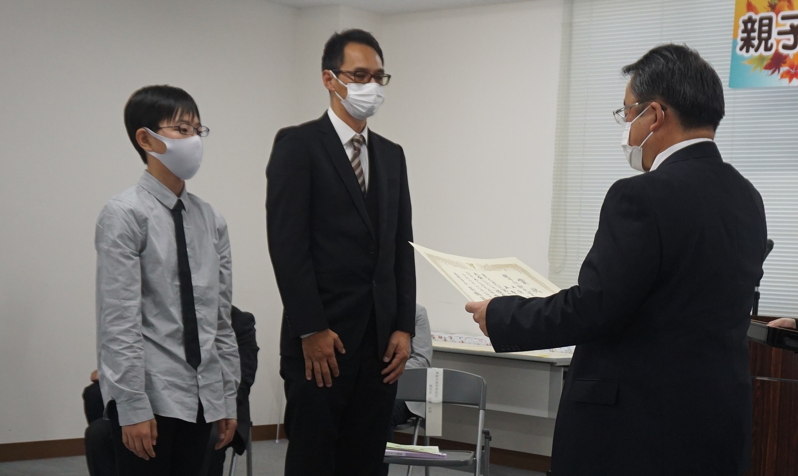並んで立った男性と男の子の前に立った、スーツを着た男性が賞状を読み上げている表彰式の様子を写した写真