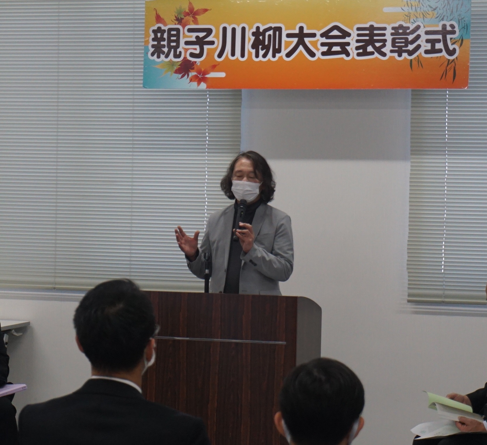 親子川柳大会表彰式と書かれた横断幕の下で、グレーのスーツを着た志水実行委員長兼審査委員長が演台に立ち、マイクを持って出席者に向かって話している写真