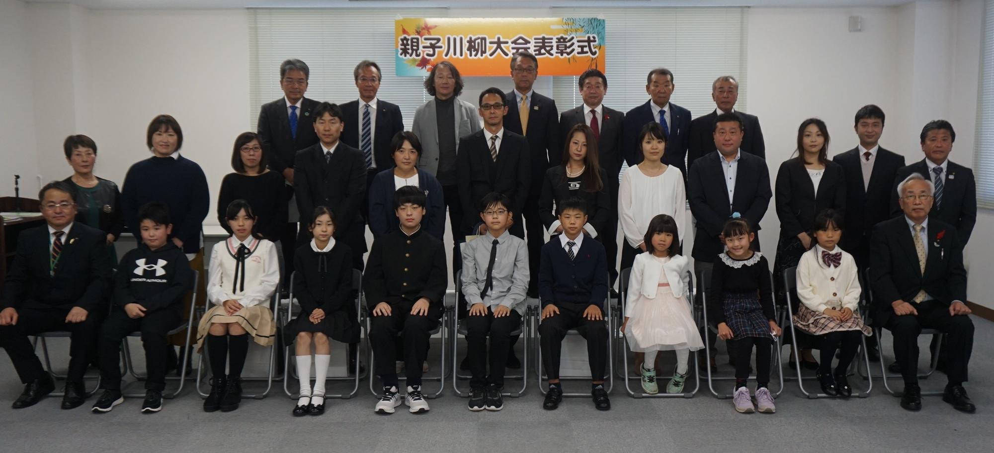 親子川柳大会表彰式と書かれた横断幕の前で、受賞者の親子や関係者が前後3列に並んで写した記念写真