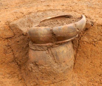 一部が破損した茶色い大きな土器が、地層に埋もれた状態で出土している発掘現場の写真