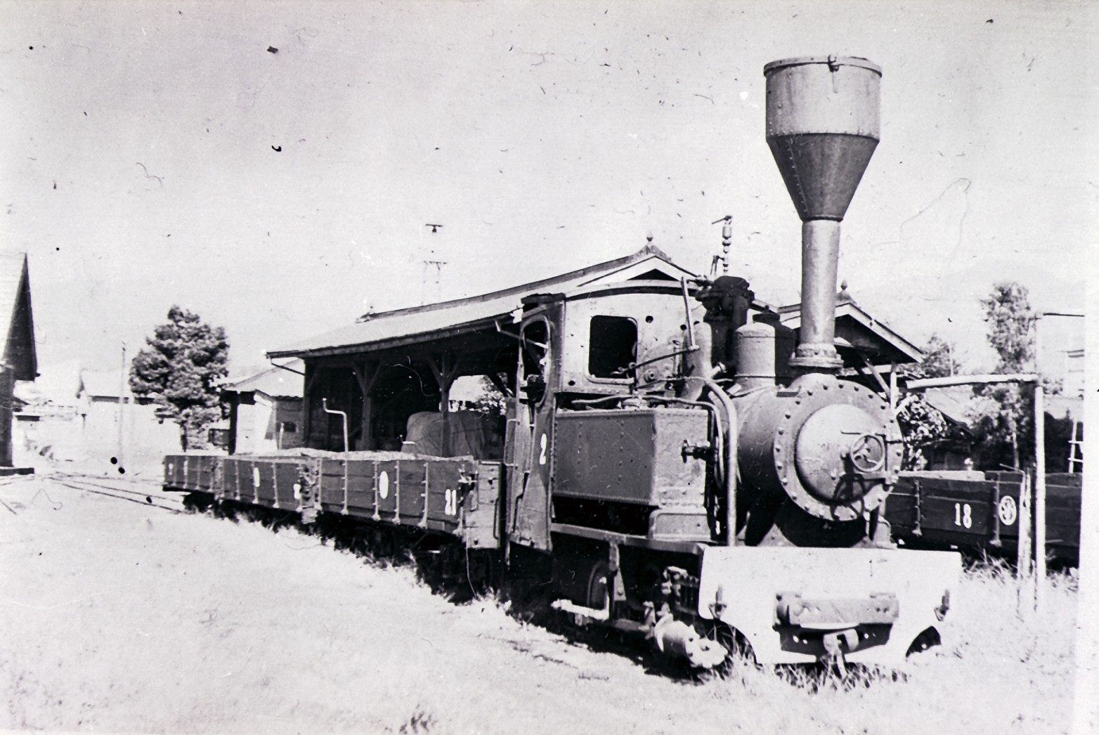 駅舎の前に停車している貨車を連結した、大きな煙突の蒸気機関車を写したモノクロ写真