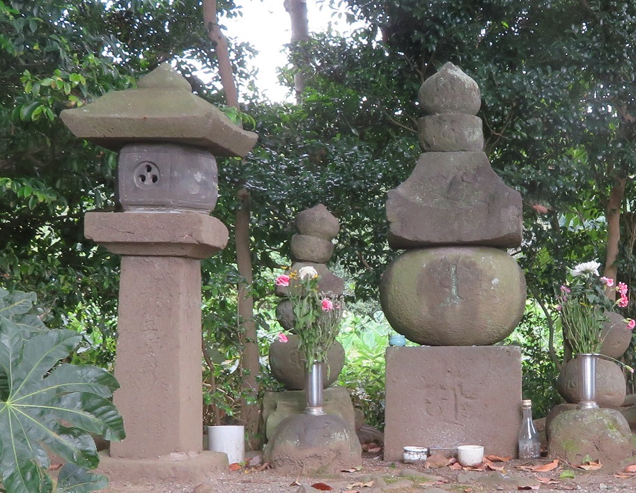 草木の中に、花や湯呑み、瓶などが供えられている石灯籠と石塔が複数並んでいる写真