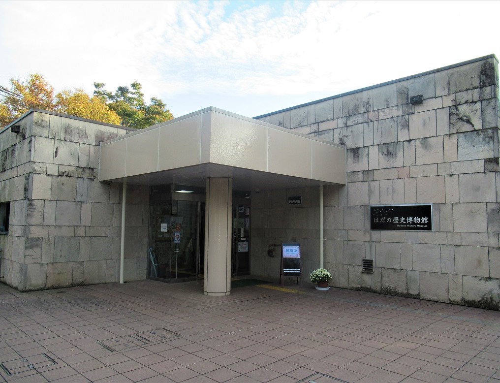 淡い茶色やグレーの大きさが違う四角形の石を組み合わせた外壁の建物の中央に、四角い厚みがある雨除けが付いたエントランスがある、秦野歴史博物館の入り口外観写真