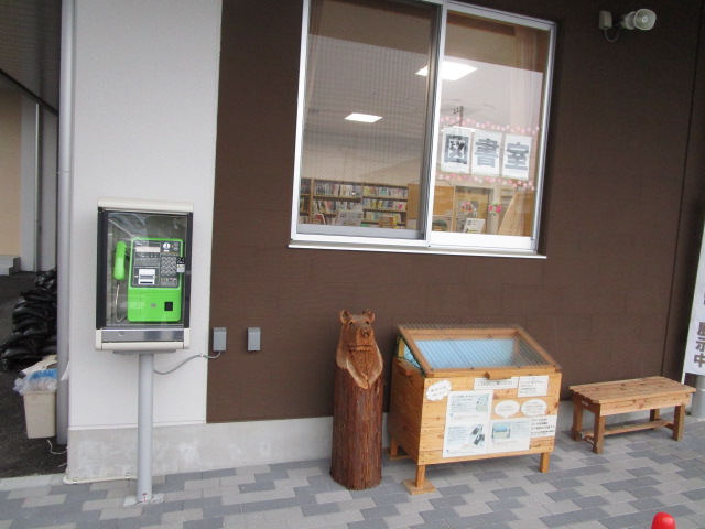 茶色の壁の正面玄関に緑色の公衆電話、木製のキエーロが置かれている写真