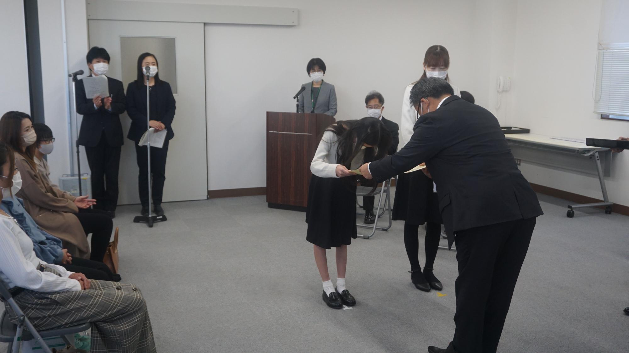 複数の関係者や参加者が見守っている中で、スーツを着た男性が入賞者の女の子に賞状を渡している様子を写した写真