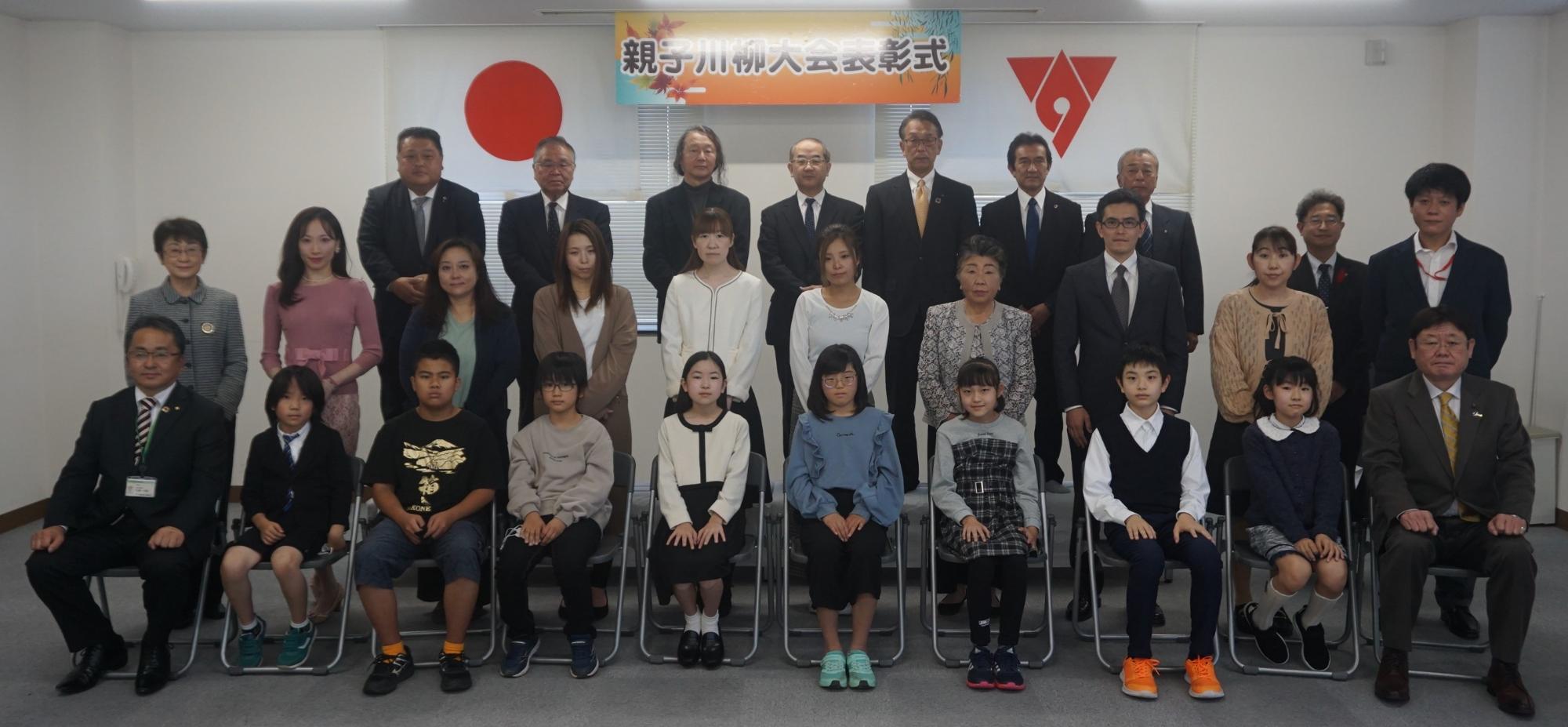 親子川柳大会表彰式と書かれた横断幕の前で、前列の椅子に入賞者の子どもたちが座り、その後ろに保護者や後援団体の方々が立ち3列に並んで写した記念写真