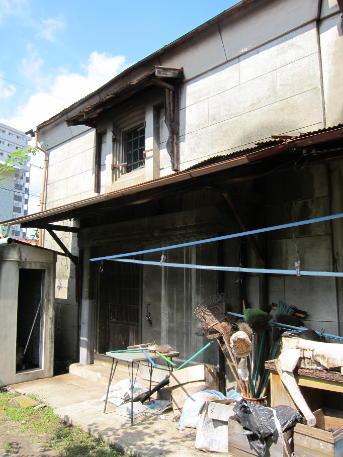 年季の入った二階建ての建物の玄関先に、たくさんの掃除用具や使われなくなった道具などが雑然と置かれている様子を写した写真