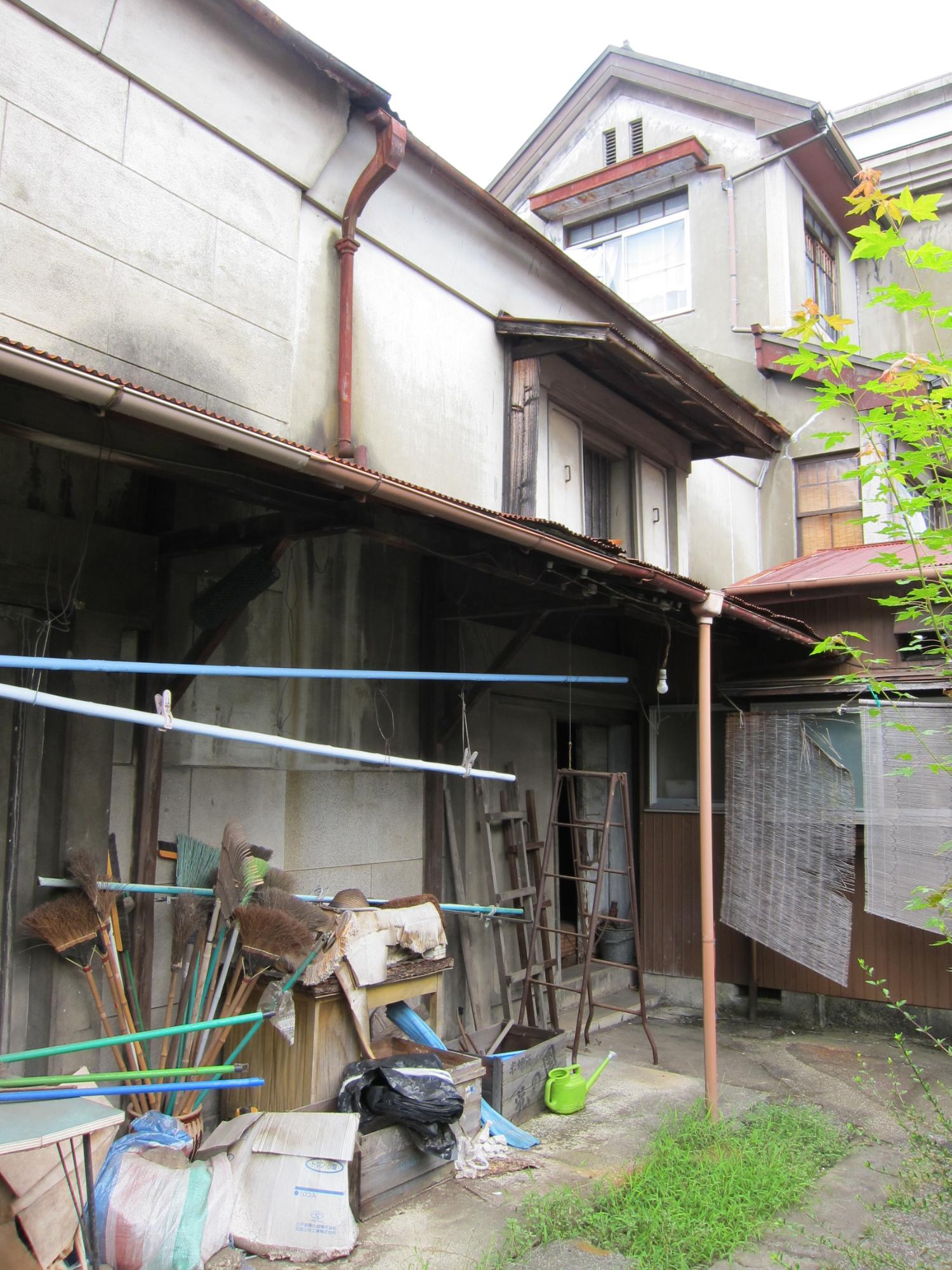 老朽化した建物に沿うように、脚立やほうきなどの清掃用具が雑然と置かれている様子を写した写真