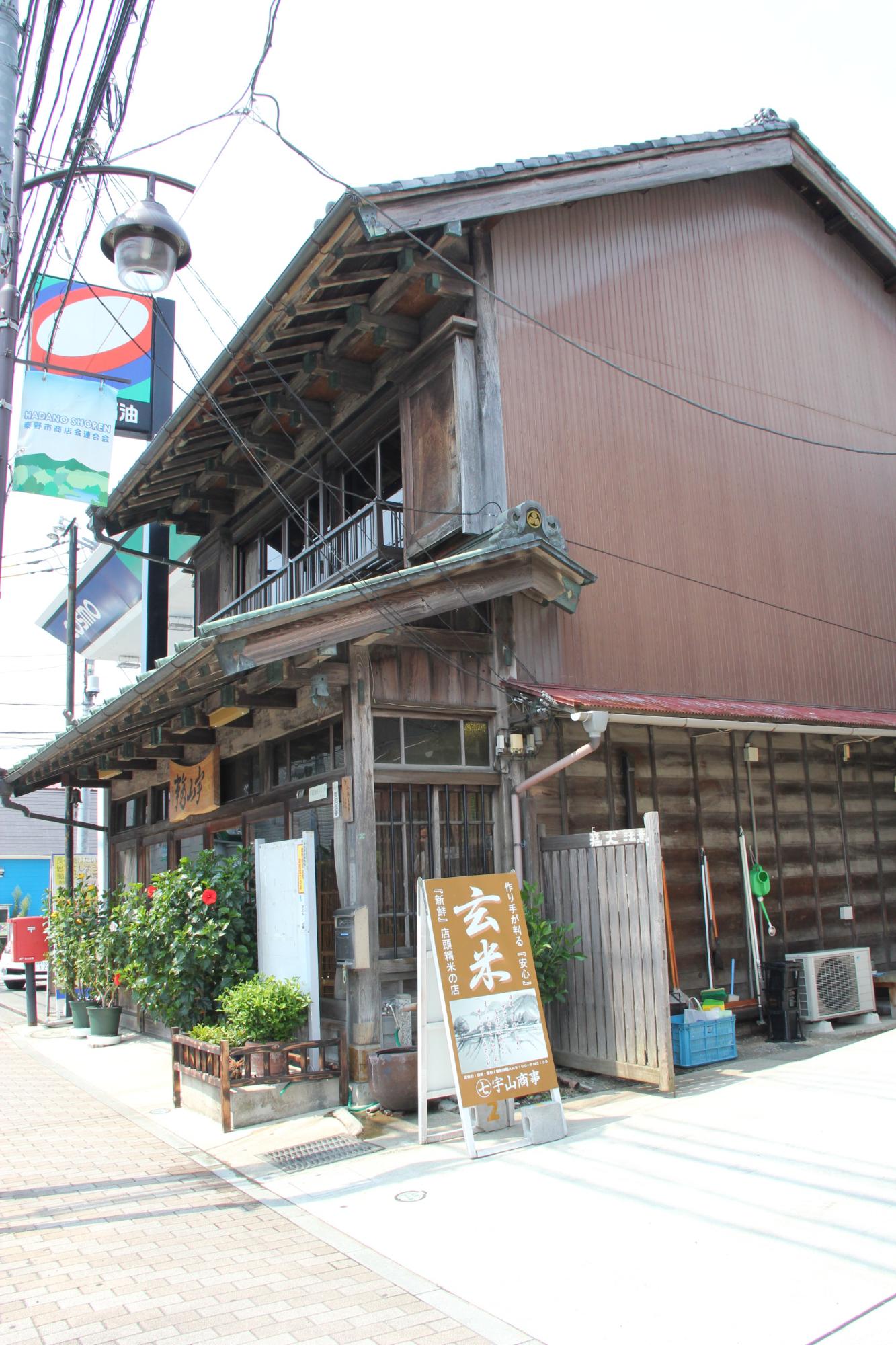 立て看板や鉢植えの植物が置かれた濃い茶色の木造の二階建ての古民家の建物を北側から写した写真