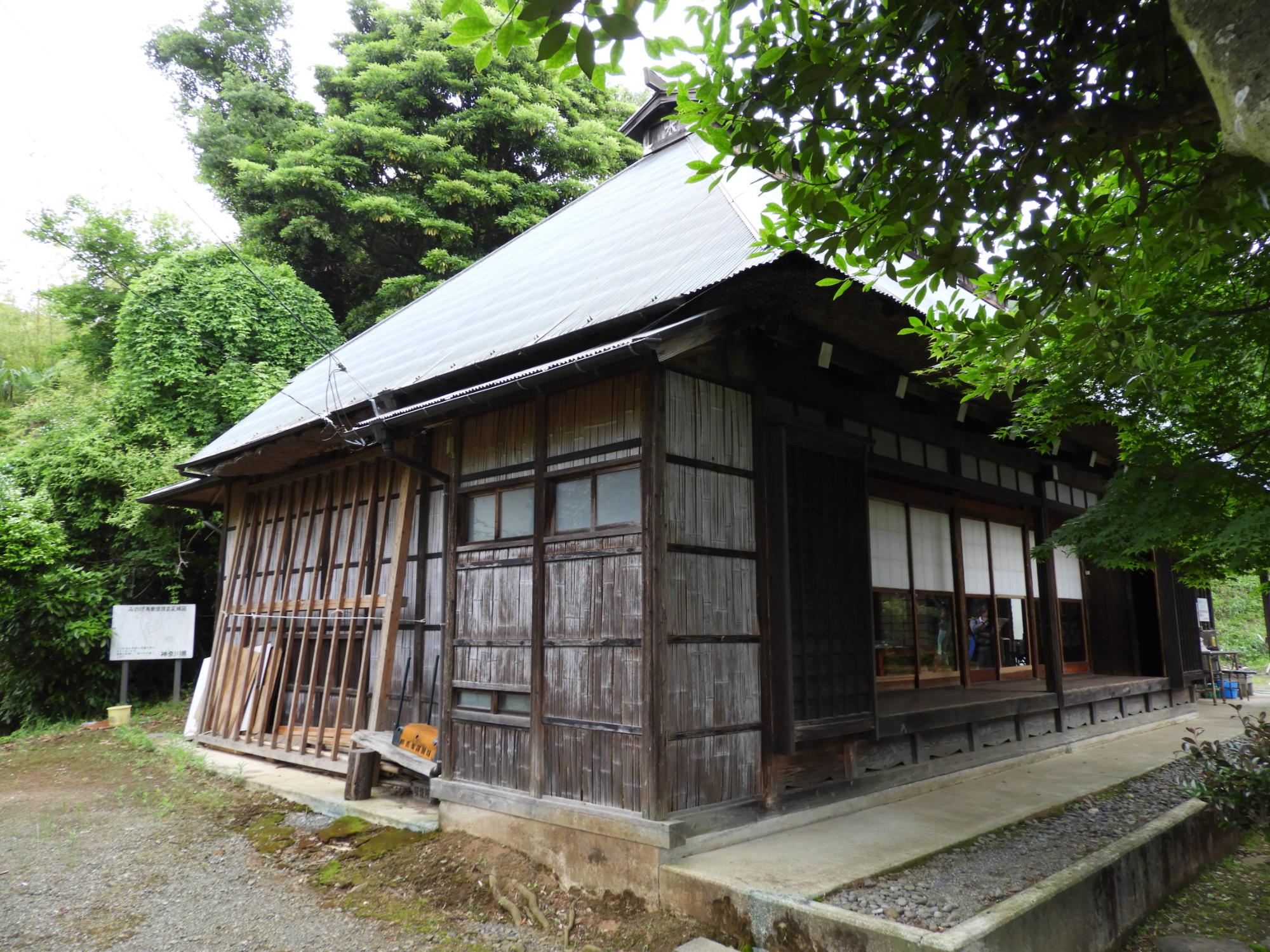 木造の家で、板張りと縦格子の外壁、縁側と障子窓、屋根に波板トタンが載った古い民家を西側から写した外観の写真