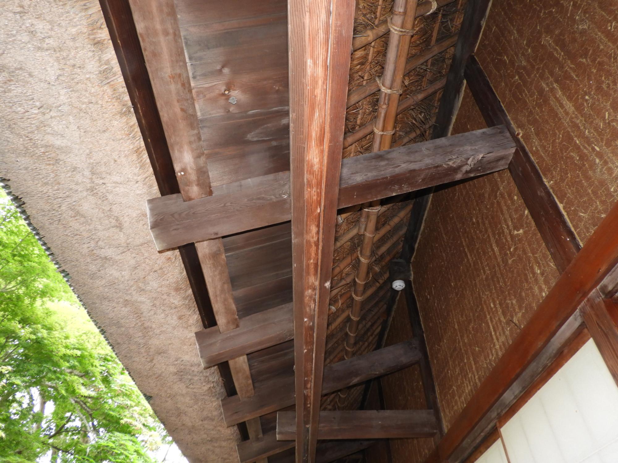 古びた濃い茶色の木製の垂木や桁と、荒い木材で組まれた軒天、そしてその奥に竹と藁の素材が見える、和風建築の軒下をアップで写した写真