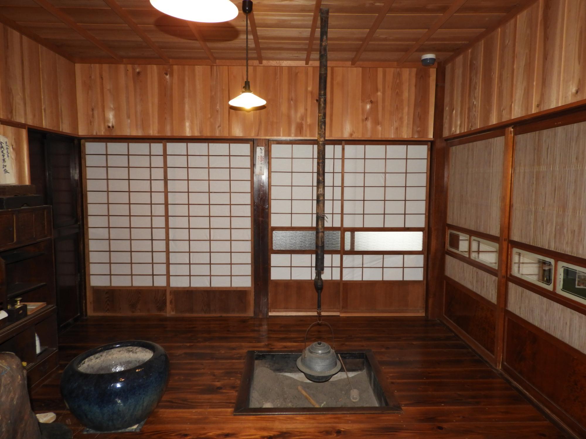 部屋の中央には四角い囲炉裏が掘られ、自在鉤に鉄瓶が吊るされ、床と壁と天井は全て明るい茶色の木材で造られた古民家の室内を正面から撮影した写真