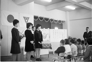 日の丸や学校の校章旗が掲げられた屋内のステージで、3人の女性が立ち、その前にいる子供たちが椅子に座っている、ひろはた幼稚園入園式の様子のモノクロ写真