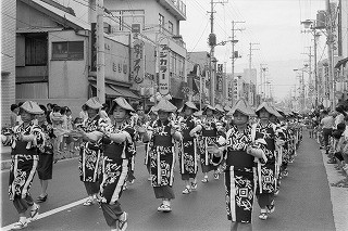 列に並んで同じ着物と笠を身に着けて踊っているたくさんの女性が、沿道にいる多くの観衆の前でパレードをしている様子のモノクロ写真
