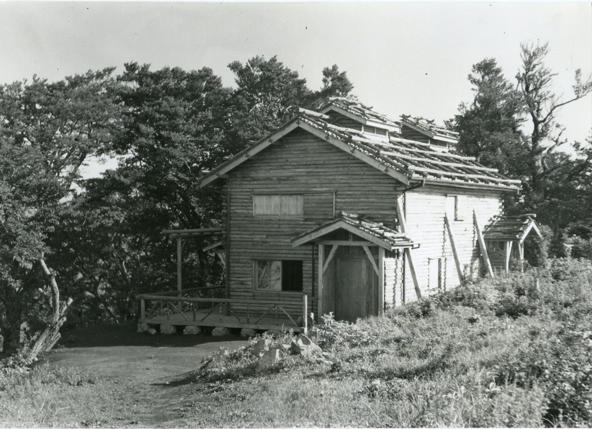 周囲を木々に囲まれた建っている板張りの壁の初代尊仏小屋のモノクロ写真