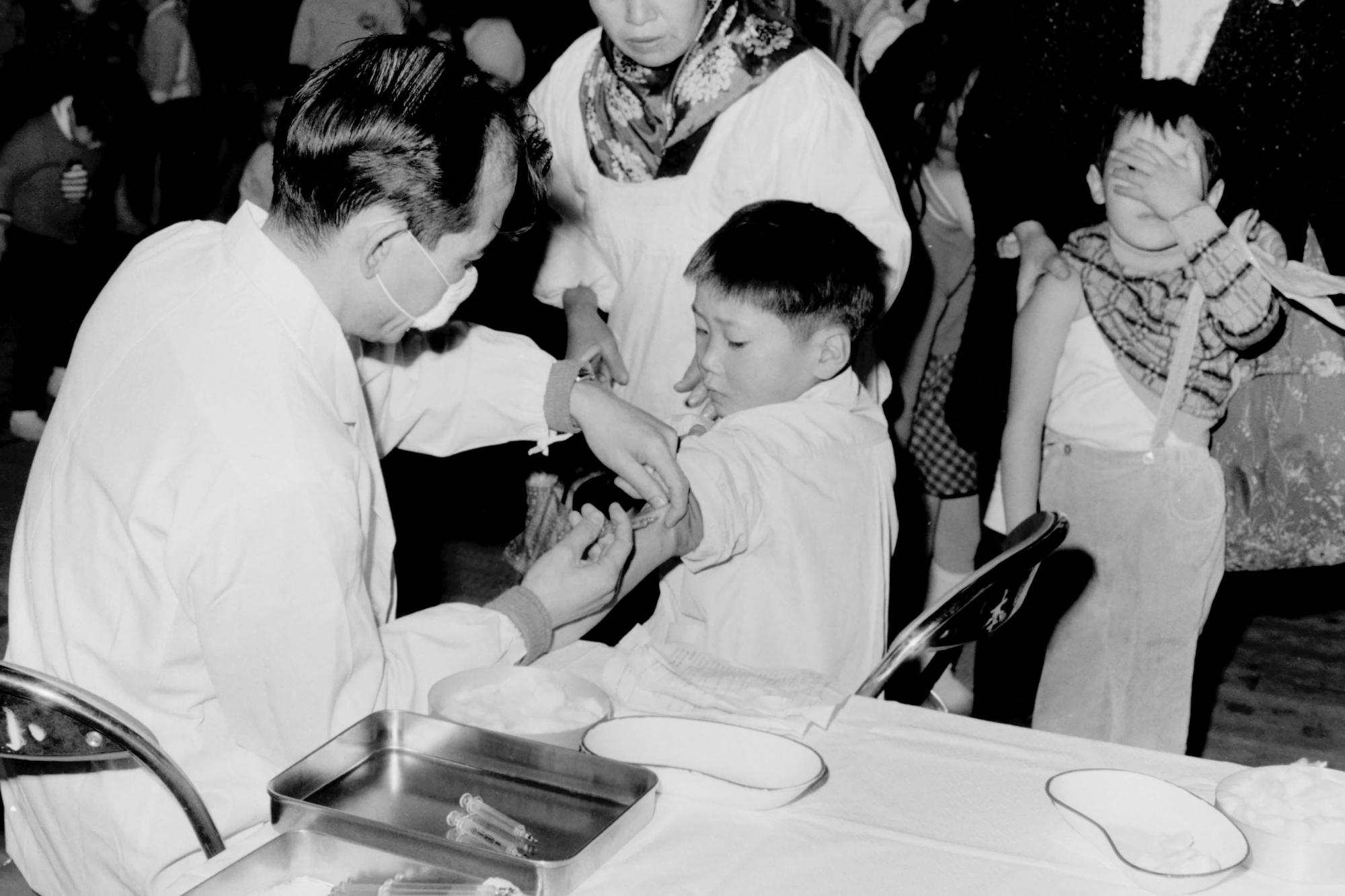 白衣とマスクを着用した医師が、椅子に座った子どもに注射をしており、列に並んだ子どもが顔を手で覆っている、予防接種の様子の写真