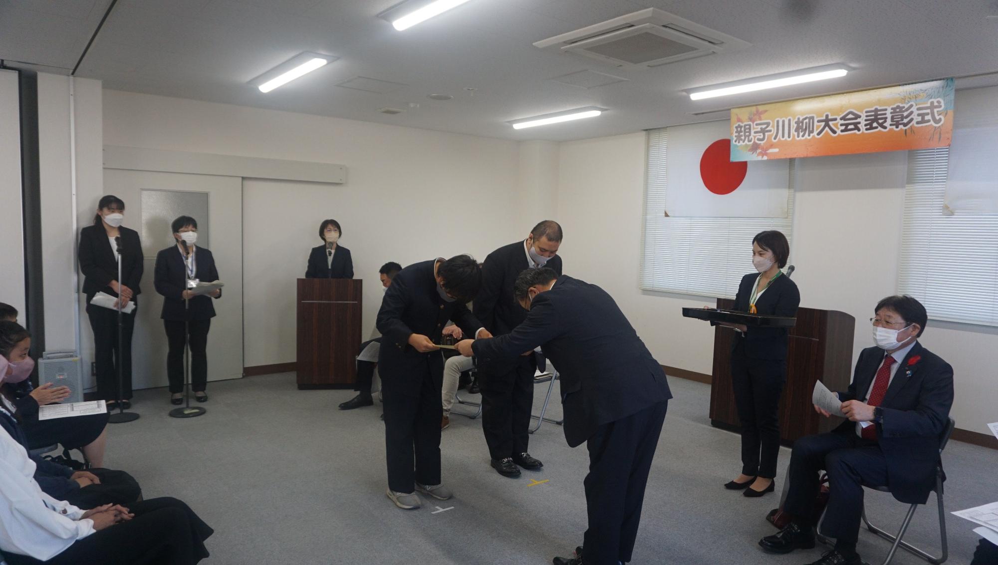 複数の関係者や参加者が見守っている中で、スーツを着た男性が入賞者の男子生徒に賞状を渡している様子を写した写真