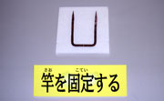 「竿を固定する」と書かれた札が置かれ、細い棒がU字型に曲がっており、タバコの葉を乾燥させるための竹を固定する道具の写真