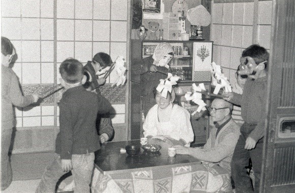 複数の子供たちがお面をつけて、炬燵に座った男女の周りを歩いている、賑やかなあくまっぱらいの風景を撮影した白黒写真