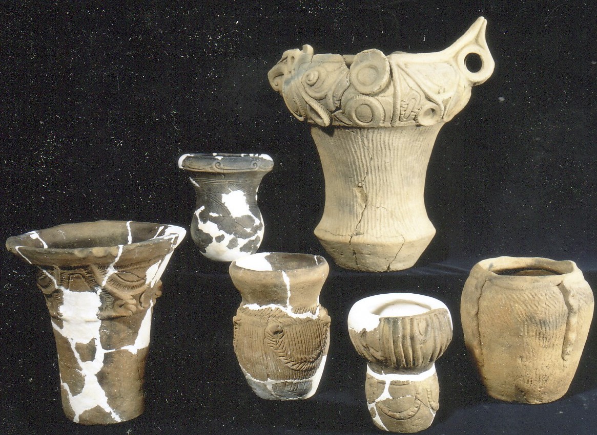 異なる時代や様式を持つ複数の土器や陶器が、暗い背景の展示台に並べられており、中には一部が白く彩色されているものや、表面に模様が施されているものがある考古学的な展示物の写真