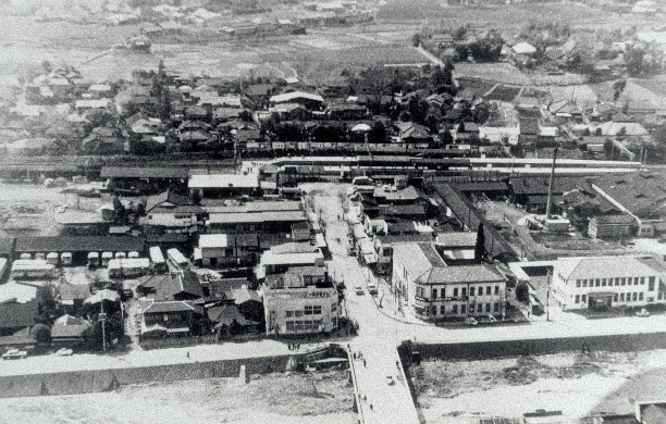 道路を挟んで多数の建物が並び、手前には橋、奥には駅のホームや線路が見え、広大な畑や田園地帯に隣接している街並みを空から撮影した白黒写真