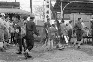 屋外の屋根付きの休憩所の前に、兵隊の服装をした男性たちや子どもなど多くの人が集まっている様子が写っている渋沢駅前の白黒写真