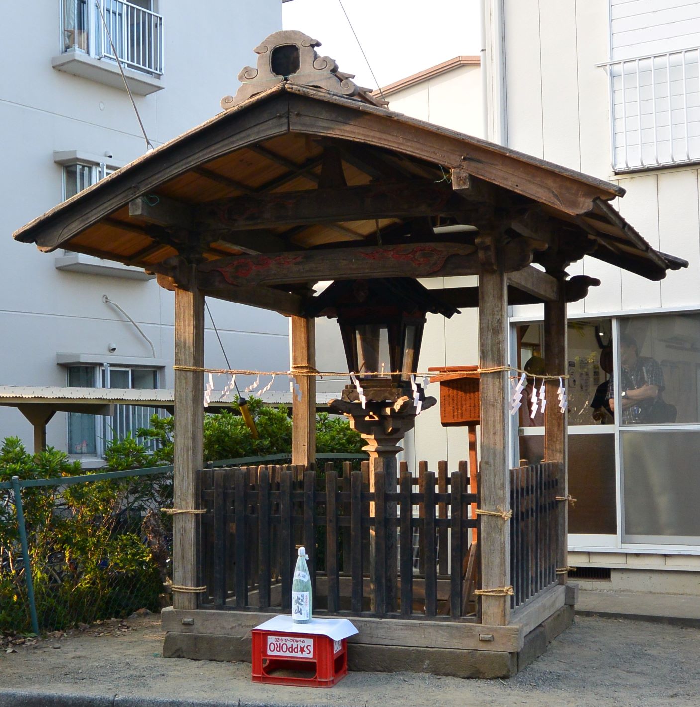 木製の小さな屋根と柵で囲まれた中央に、白く細長い紙が吊り下げられた常夜灯が設置されており、手前にはお酒の瓶が置かれている大山灯籠の風景を撮影した写真