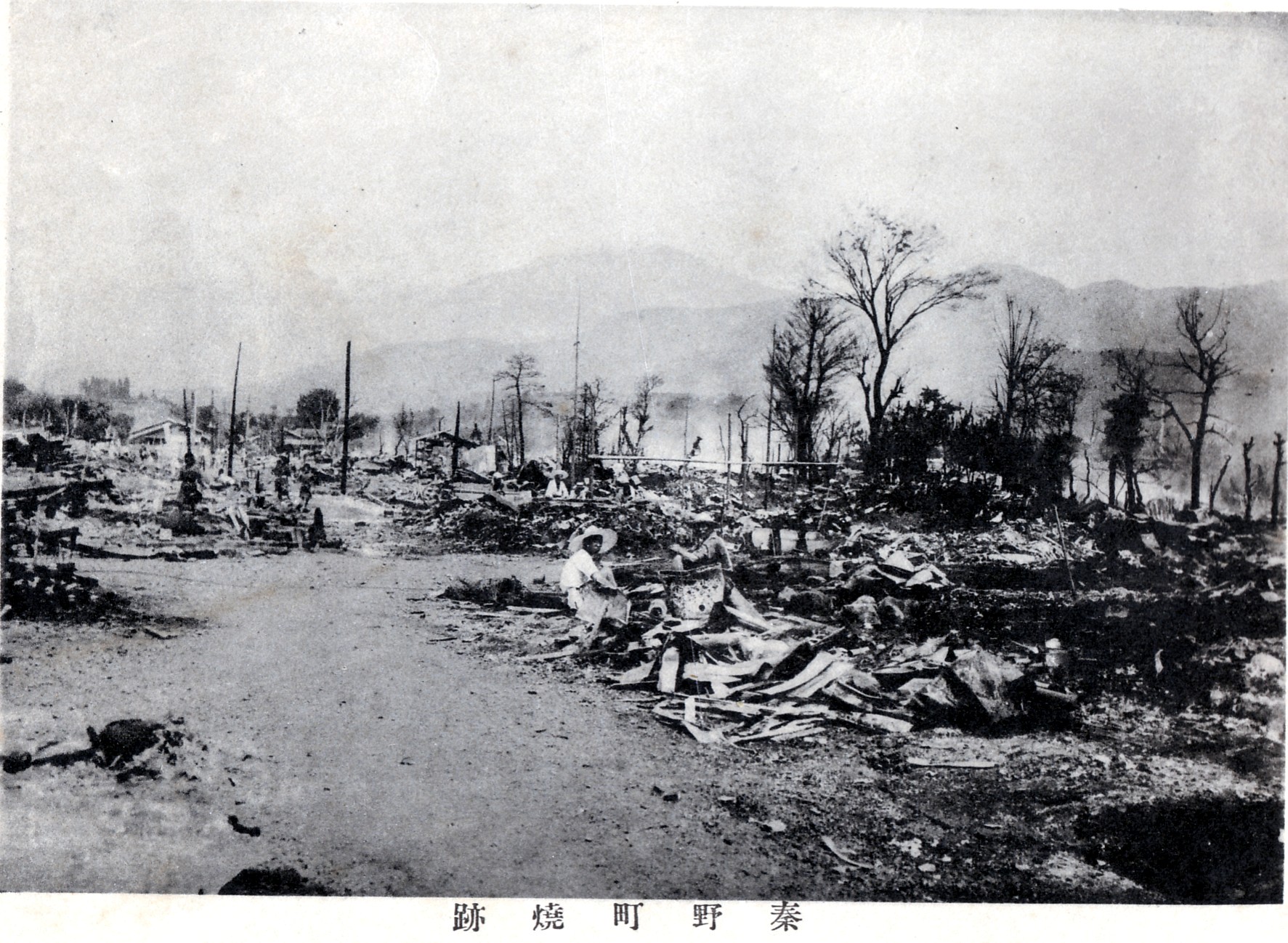 建物がすべて焼け、焼け焦げた木々や瓦礫が広がる広い土地に数人の人物がおり、煙に霞んだ先に山々の輪郭が見える白黒写真